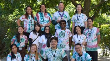 Team wearing matching tie-dye shirts poses outside under a tree, representing group unity and spirit at UC Davis.