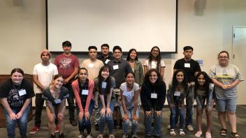 Group of students posing in two rows indoors, smiling for a group photo in front of a projector screen.