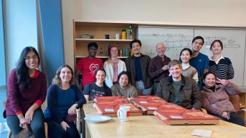 Lab members gather around a table with pizza boxes in a room with whiteboards and shelves, enjoying a casual meeting.
