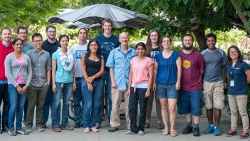 Lab group stands outside under trees and umbrellas. Diverse group smiles together during a casual outdoor gathering.