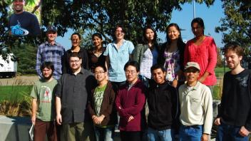 Lab group poses on sunny day near trees and benches, smiling in two rows on grass, representing collaborative research team.