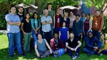 Group of scientists and students smiling outdoors in front of leafy trees, wearing casual clothing in sunny spring setting. 