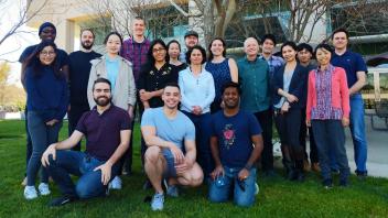 Group of lab members smiles outside on green grass, posing together in front of a building with trees and windows.