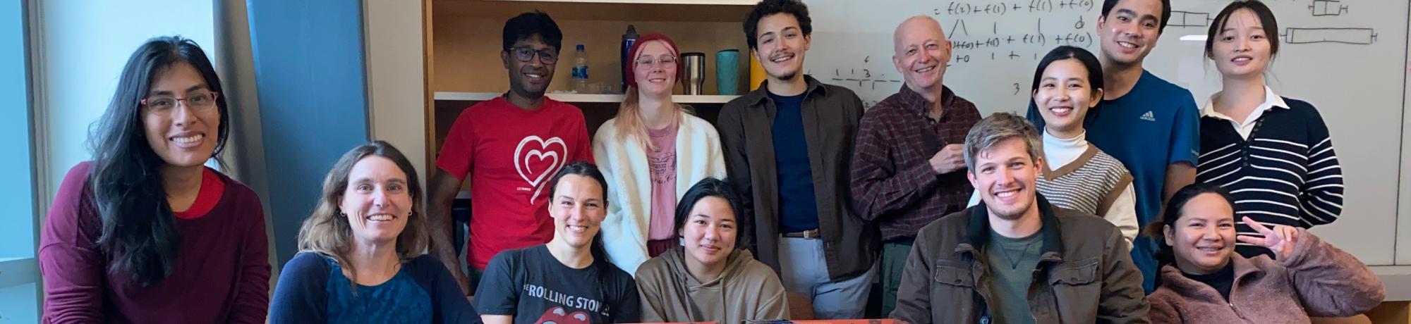 Lab members gather around a table with pizza boxes in a room with whiteboards and shelves, enjoying a casual meeting.